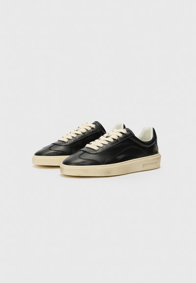 Zapatillas de cuero negro con cordones crema, con una puntera texturizada y suela de goma color blanco sucio. El lado presenta un sutil detalle en dorado.