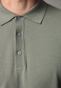 Polo cu mânecă scurtă, de culoare verde deschis, din tricot, având un guler și trei nasturi gri, cu un finisaj de țesătură texturată.