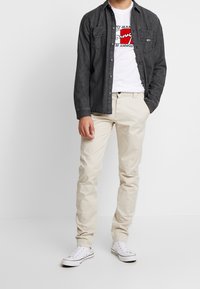 Mörk denimskjorta med knappar över en vit t-shirt med grafiskt tryck och röda accenter, i kombination med beiga chinos och vita sneakers.