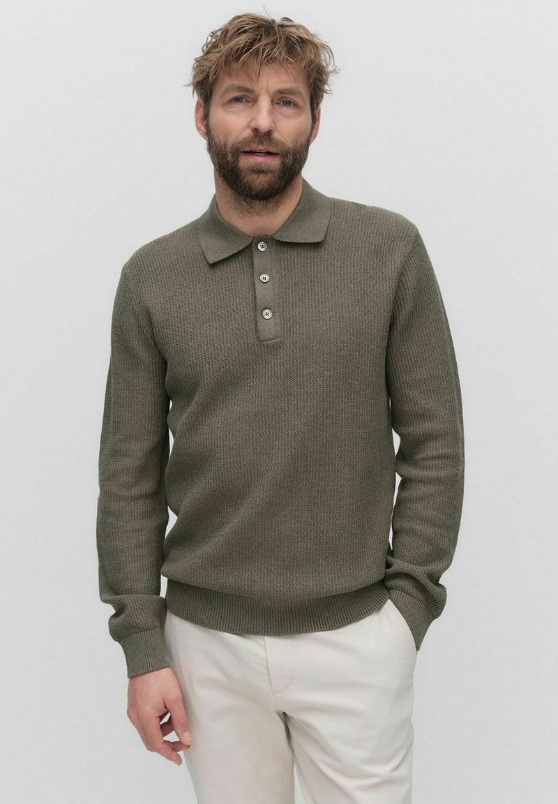 Olivgrön långärmad pikétröja med texturerad stickning, tvåknapplås och ribbad nederkant. Bärs över ljusa byxor.