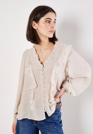Light beige blouse with ruffled detailing, button front, and long puffed sleeves. Fabric appears lightweight and textured. Paired with blue jeans.