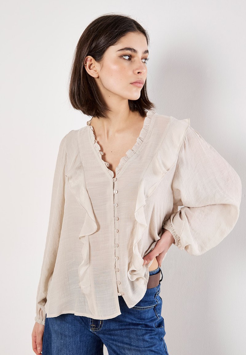 Blusa beige chiaro con dettagli arruffati, chiusura frontale a bottoni e maniche lunghe a sbuffo. Il tessuto sembra leggero e testurizzato. Abbinata a jeans blu.