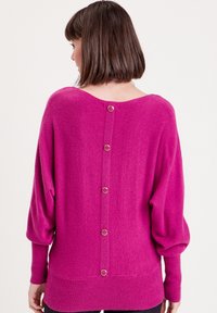 Pull fuchsia à la coupe décontractée, avec un dos en forme de scoop, un ourlet côtelé et des accents de boutons métalliques le long de la colonne vertébrale. Texture douce.