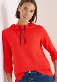 Punainen huppari, jossa on röyhelöhuppu ja tummansiniset nyörit, kolme neljänneksen hihat ja rento istuvuus. Yhdistetty sinisiin farkkuihin.