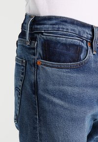 Primer plano de la zona de la cintura de unos vaqueros de mezclilla azul con bolsillos delanteros y remaches de cobre, usados con una camisa blanca metida.