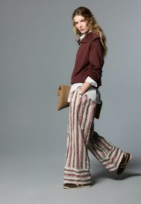 Bordeauxrode trui met capuchon, witte overhemd met kraag eronder, en beige wijde broek met rode strepen. Bruine schoenen en een tas met structuur.