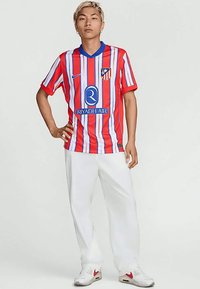 Noor inimene seisab käed puusadel, seljas punase ja valge triibulise Atlético Madridi jalgpallipluusi, valgete pükste ja valgete tossudega.