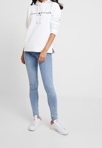 Biała bluza z kapturem z czarnym logo "Tommy Hilfiger", zestawiona z jasnoniebieskimi obcisłymi dżinsami i białymi sneakerami z pomarańczowymi akcentami.