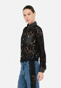 Camicetta nera in pizzo con dettagli in denim. Stile button-up, maniche lunghe e motivo floreale. Presenta una tasca sul petto e bottoni decorativi.