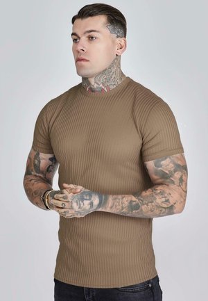 Mand med fulde ærmetatoveringer, hals- og hovedtatoveringer, iført en brun ribbet kortærmet skjorte, sammenfoldede hænder, vendt mod venstre mod en ensfarvet baggrund.