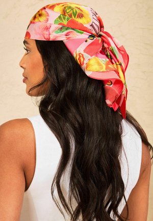 Woman with long dark hair wearing a white sleeveless top and a pink floral scarf tied around her head, viewed from the side and back.