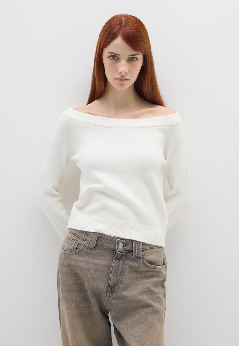 Beige, schulterfreier Pullover aus weichem Stoff mit geripptem Saum. Kombiniert mit hellbraunen, ausgewaschenen Jeans mit Fünf-Taschen-Design.