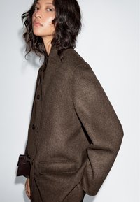 Brown, textured wool coat with a high collar, double-button closure, and side pockets, featuring a relaxed fit and smooth fabric.