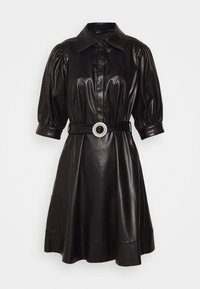 Robe chemise en cuir noir avec des manches bouffantes, une taille ceinturée mettant en valeur une boucle circulaire ornée de bijoux, et une ourlet évasé.