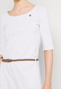 Jasnoszara sukienka z długim rękawem i szerokim dekoltem, z małym srebrnym akcentem i plecionym brązowym paskiem w talii. Miękka tekstura materiału.