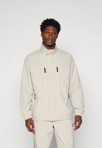 Giacca zip-up beige chiaro con colletto alto, polsini elastici e due tasche frontali. Caratterizzata da maniglie della cerniera nere e un tessuto liscio e strutturato.