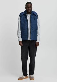 Jūras spēku krāsas pūcija vestē ar kapuci, ar priekšējo rāvējslēdzēju un quiltotām sekcijām, valkāta virs pelēkā sporta krekla, kombinēta ar tumšajām biksēm un sporta apaviem.