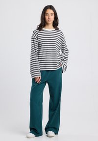 Vrouw die staat en een zwart-wit gestreepte blouse met lange mouwen, teal wijde broek en witte sneakers draagt, tegen een effen achtergrond.
