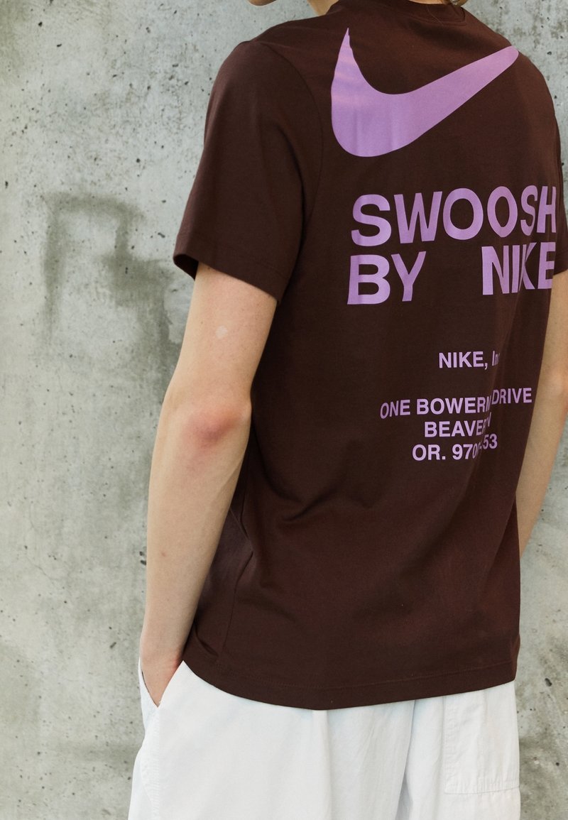Brązowy bawełnowy T-shirt z dużym różowym logo swoosh Nike oraz dużym fioletowym napisem "SWOOSH BY NIKE" na plecach.