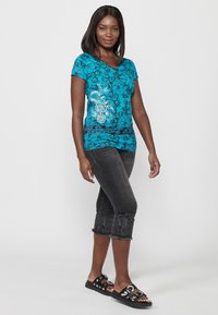 T-shirt turquoise à fleurs en V avec des manches courtes, associé à un jean raccourci gris foncé avec des bords brodés et des sandales noires à enfiler.