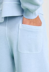 Un pantalon de survêtement bleu clair en coton doux, avec une grande poche latérale et un logo de marque en relief, ainsi qu'une taille et des poignets côtelés.