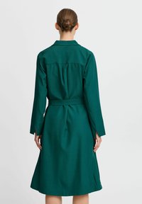 Grünes Hemdkleid mit langen Ärmeln, einem Kragen, einem taillierten Gürtel und einem schwingenden Rock, aus glattem Stoff mit minimaler Textur.