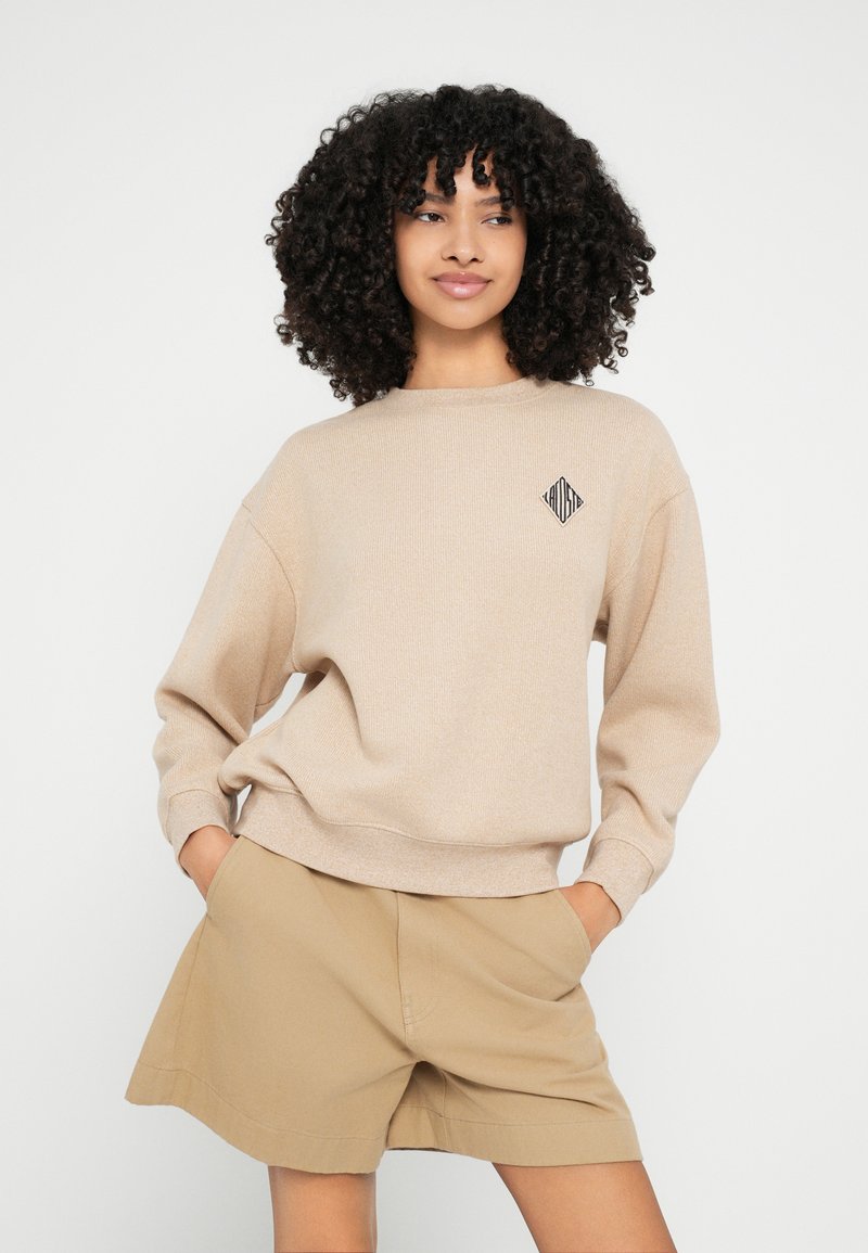 Beige gestructureerd sweatshirt met een ronde hals en een zwart geometrisch logo op de borst, gecombineerd met beige shorts.