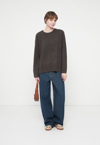 Mörkbrun stickad tröja med rund hals, kombinerad med lösa blå jeans. Modellen håller en liten brun handväska och har på sig beige skor med slipp på.
