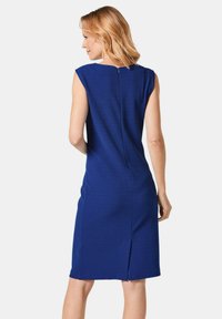 Blaues knielanges Kleid mit taillierter Form, ohne Ärmel, mit Reißverschluss am Rücken und einem kleinen Schlitz am Saum. Texturiertes Material verleiht Tiefe.