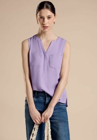 Top lavanda senza maniche con scollatura a V, due tasche frontali e un design fluido; indossato con jeans blu scuro e con in mano una maniglia a corda.