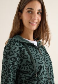 Felpa verde con zip caratterizzata da un motivo a stampa leopardato nero, tessuto testurizzato e cappuccio con cordino. Maglietta bianca visibile sotto.