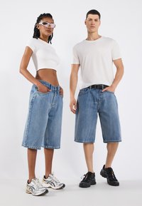 Kék farmer rövidnadrágok, lazán illeszkedő fazonnal, két modellen. Az egyik egy fehér crop topot és napszemüveget visel; a másik sima fehér pólót hord.
