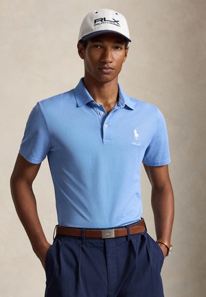 Jeune homme portant un polo bleu, un pantalon foncé avec une ceinture marron, une casquette blanche avec le logo RLX et une montre-bracelet, debout les mains dans les poches.