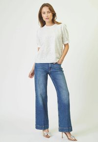Blusa bianca ricamata con maniche corte, abbinata a jeans in denim blu a gamba larga. Accentuata da sandali con tacco di colore chiaro con fiocchi.