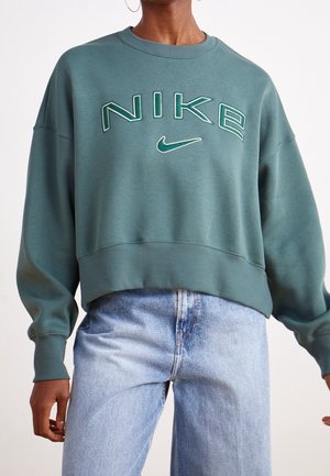 Personne portant un sweat Nike turquoise avec un grand logo et un jean en denim bleu clair, montrée du cou aux hanches sur un fond uni.