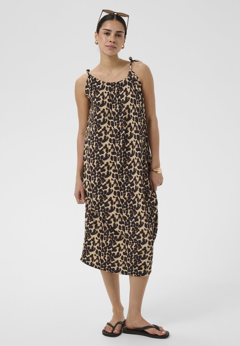 Femme portant une robe midi sans manches imprimée léopard, des tongs noires, des boucles d’oreilles créoles dorées, des bracelets et des lunettes de soleil posées sur sa tête.