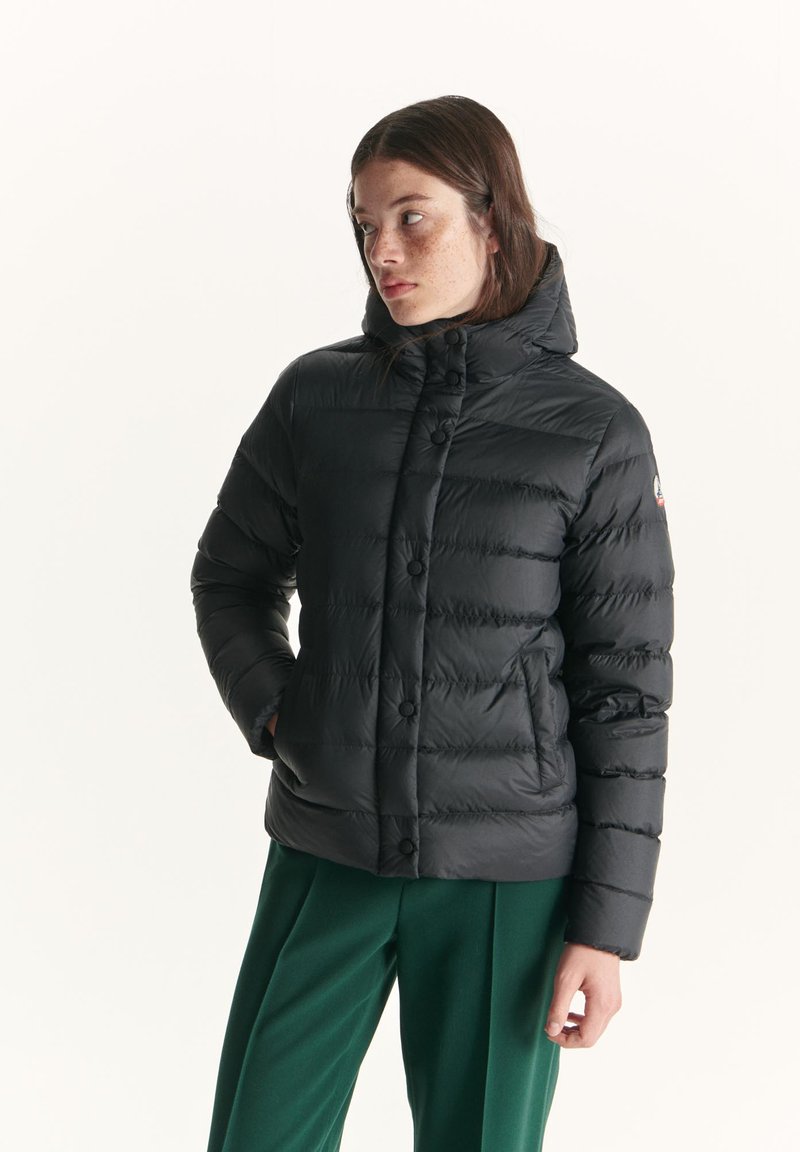 Giacca a vento nera con colletto alto, bottoni frontali e trapuntatura orizzontale; indossata con pantaloni verdi. Texture liscia e lucida.