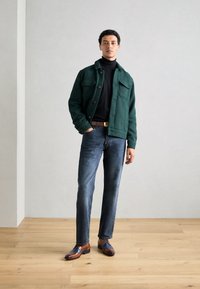 Giacca verde strutturata con due tasche sul petto, dolcevita nero, jeans blu e scarpe eleganti marroni e blu, in piedi su un pavimento in legno.
