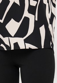 Svart topp med et kremfarget abstrakt mønster, laget av lett stoff med en lett teksturert overflate. Kombinert med svarte tettsittende bukser.