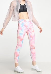 Lekka, przezroczysta różowa kurtka na czarnym staniku sportowym. Legginsy w wzory z pastelowymi kształtami geometrycznymi w odcieniach różu, niebieskiego i białego. Białe sneakersy.