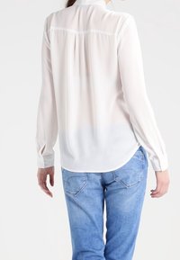 Blusa bianca a maniche lunghe in tessuto leggero con orlo curvo, caratterizzata da un dettaglio a yoke e polsini con bottoni, abbinata a jeans blu.