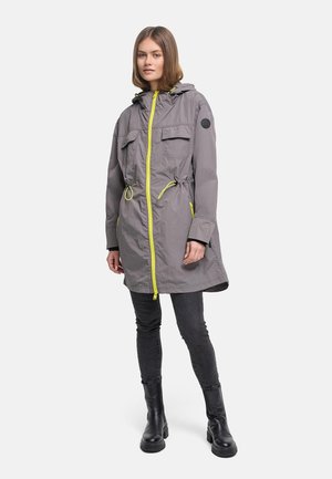 Kvinne iført knelengde grå hettejakke med gul glidelås og snorer, svarte jeans og svarte støvler, står mot hvit bakgrunn.