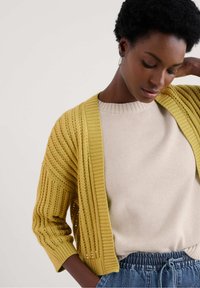 Gelber, offen gestrickter Cardigan mit gerippten Kanten, über einem hellbeigen Pullover getragen. Das Outfit hat einen lockeren Schnitt und strukturierte Designs.