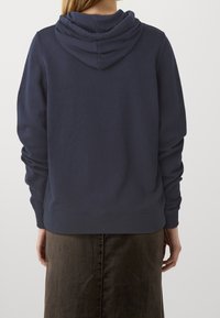 Námořnický modrý sweatshirt s uvolněným střihem, s dvojitě vrstvenou kapucí, žebrovanými manžetami a lemem. Tkanina se zdá být měkká a texturovaná.