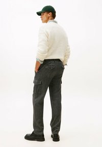 Hombre con gorra verde, suéter crema sobre camisa a rayas, pantalones cargo grises y zapatos negros, de pie con las manos en los bolsillos, mirando de lado.