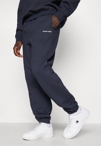 Μπλε φόρμες με ελαστικούς καρπούς, με λογότυπο σε λευκό χρώμα στην αριστερή τσέπη. Συνδυασμένες με λευκά αθλητικά παπούτσια.