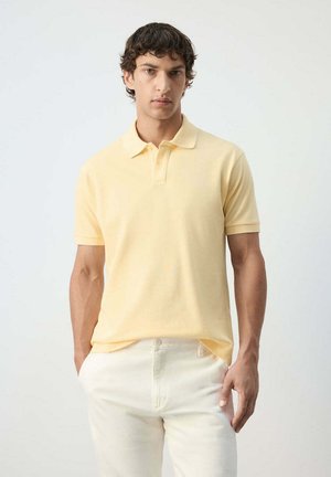 Homme aux cheveux bouclés foncés, portant un polo jaune pâle à manches courtes et un pantalon blanc cassé, debout les mains dans les poches.
