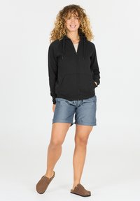 Hoodie zippé noir avec une poche avant, associé à un short bleu clair. Le modèle porte des chaussures slip-on marron, se tenant devant un fond blanc.