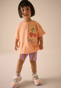 Ljusorange t-shirt med en körsbärsgrafik och rutigt mönster, kombinerad med lila shorts med gula ränder. Flerfärgade sneakers.
