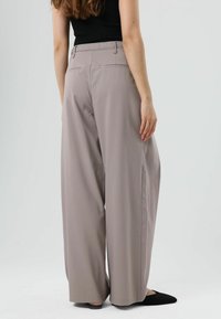 Wijde broekspijpen in lichtgrijze stof. Heeft een gladde textuur met achterzakken en een getailleerde pasvorm. Draagt zwarte schoenen.